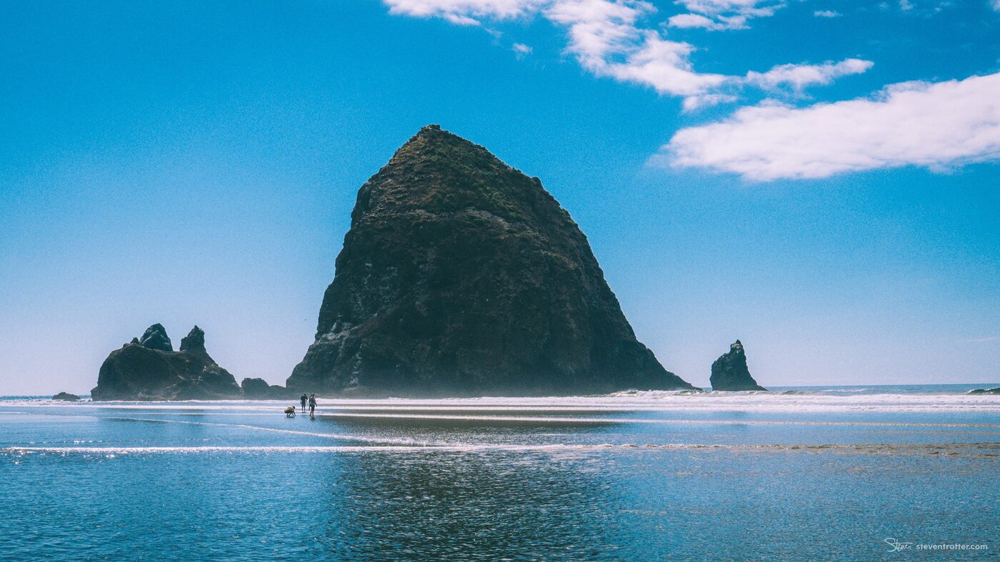 Haystack Rock at Cannon Beach under a blue sky, two people and a dog walking the wet sand below.